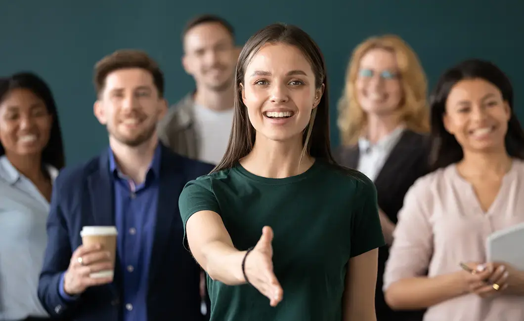 Smiling young woman extending her hand for a handshake, standing in front of a diverse group of colleagues, all looking welcoming and friendly.